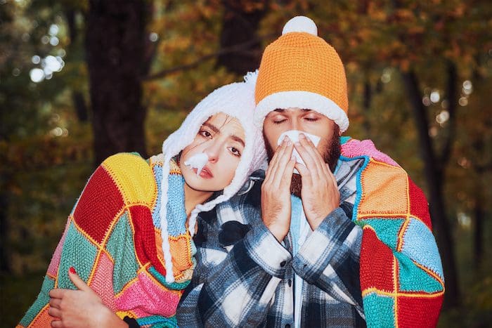 A man and woman in winter clothing blowing noses.