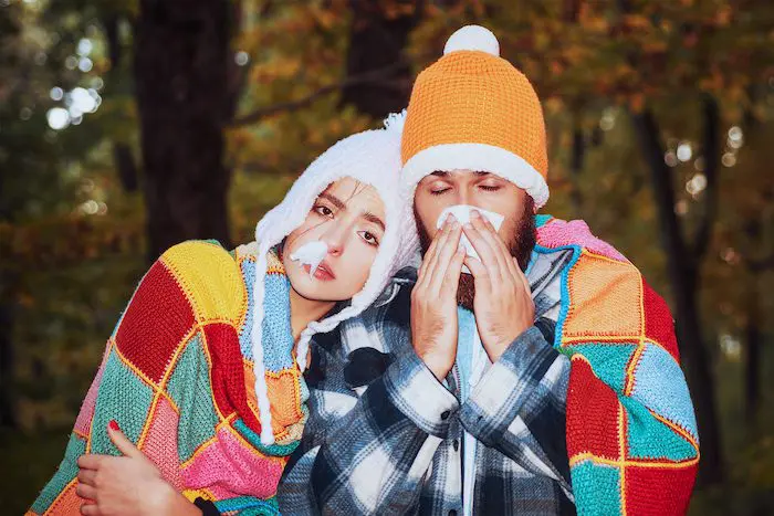 A man and woman in winter clothing blowing noses.